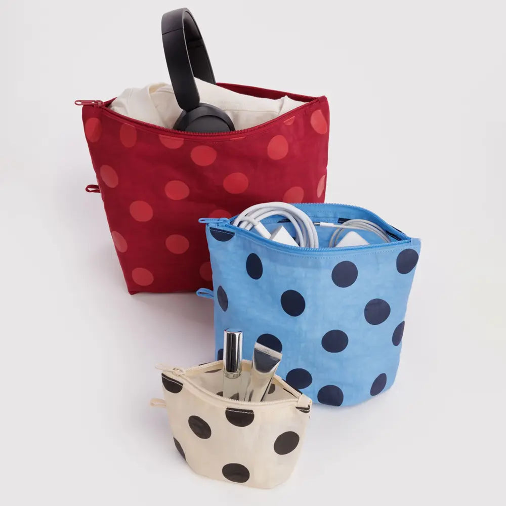 Three fabric zippered pouches with polka dots are arranged on a light gray background. The back pouch is red with red polka dots and contains a black handle and white contents; the middle pouch is light blue with dark blue polka dots and contains white cords; the front pouch is cream with dark polka dots and contains silver utensils.