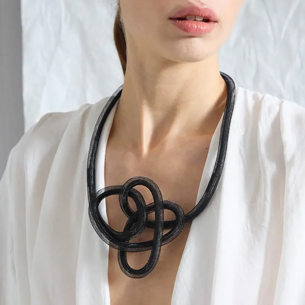 A close-up of a model wearing a chunky black mesh knot necklace over a white blouse. The necklace is from Iskin Sisters.