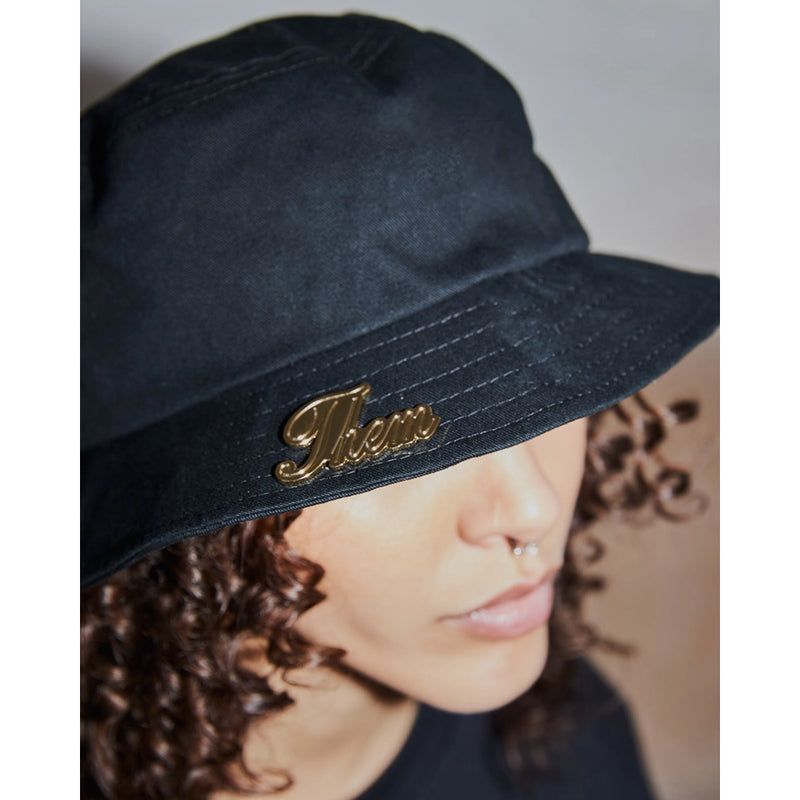 gold Us Them Pin from Gary Simmons: Public Enemy . The pin styled on a black bucket hat