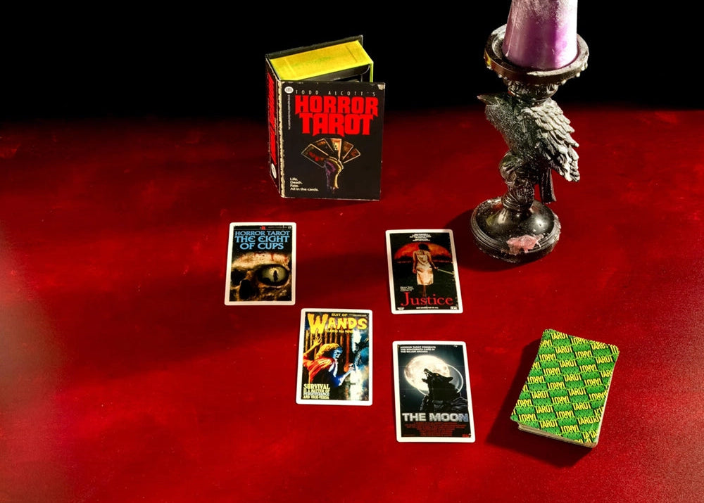 A boxed tarot deck labeled 'Horror Tarot' stands beside several illustrated tarot cards and a green patterned card stack on a red surface, with a purple candle in a metal holder against a black background.